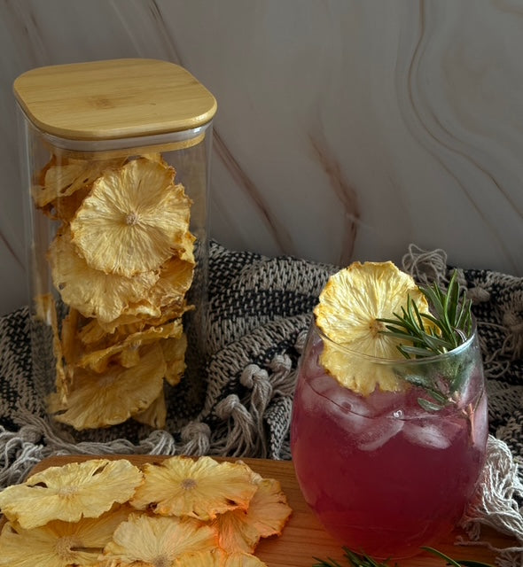 Glass jar with dried pineapple slices, a pink drink with a pineapple slice and rosemary, and a textured blanket background.